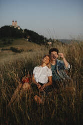 Ein junges Paar bei einem Spaziergang in der Natur in der Abenddämmerung auf dem Lande, sitzend und ruhend im langen Gras. - HPIF07159