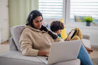 Eine indische Mutter, die einen Laptop benutzt, und ihr kleiner Sohn, der ein Tablet benutzt und zu Hause auf dem Sofa sitzt. - HPIF07350