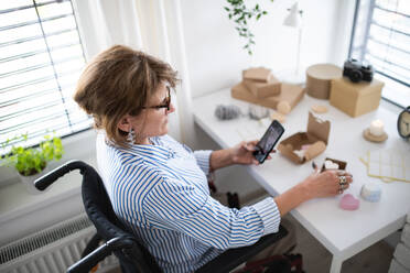 Eine behinderte reife Frau im Rollstuhl verpackt handgefertigte Produkte zu Hause, kleines Geschäftskonzept. - HPIF07380