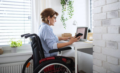 A disabled mature woman with laptop working indoors in home office, small business concept. - HPIF07381