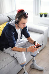 A portrait of disabled young man using smartphone indoors at home, leg prosthetic concept. - HPIF07383