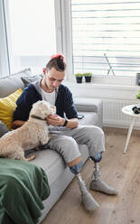 Ein Porträt eines behinderten jungen Mannes, der zu Hause mit einem Hund spielt, Konzept einer Beinprothese. - HPIF07385
