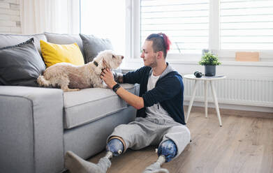 Ein Porträt eines behinderten jungen Mannes, der zu Hause mit einem Hund spielt, Konzept einer Beinprothese. - HPIF07387