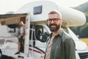 Ein Porträt eines glücklichen Mannes, der auf einem Campingplatz neben seinem Auto steht und in die Kamera schaut. Familienurlaub im Wohnwagen. - HPIF07405