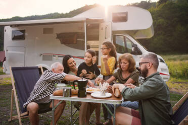 Eine Mehrgenerationen-Familie, die mit dem Auto im Freien sitzt und isst, eine Urlaubsreise mit dem Wohnwagen. - HPIF07420