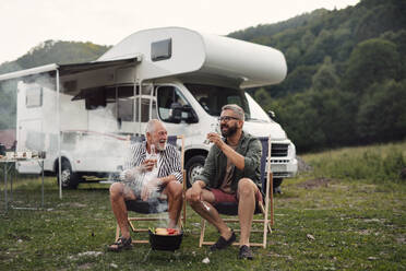 Ein reifer Mann mit seinem älteren Vater unterhält sich auf einem Campingplatz im Freien beim Grillen auf einer Wohnwagenreise. - HPIF07424