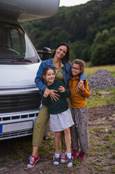 Glückliche Mutter mit Töchtern, die auf einem Campingplatz in der Abenddämmerung am Auto stehen, Familienurlaub mit Wohnwagen. - HPIF07442