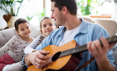 Portrait of father with small daughters sitting on sofa indoors at home, playing guitar. - HPIF07557