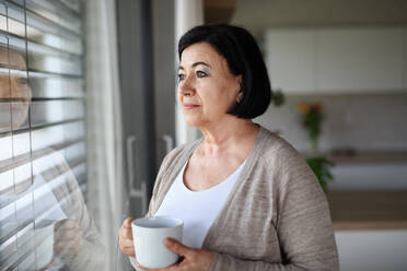 A sad senior woman standing and looking out through window indoors at home. - HPIF07579