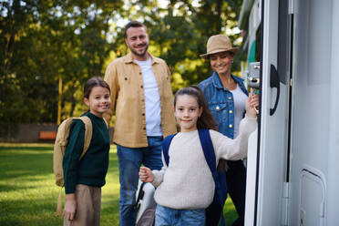 Eine glückliche junge Familie mit Gepäck, die in die Kamera schaut, mit einem Wohnmobil im Hintergrund, das im Garten steht. - HPIF07664