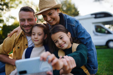 Eine fröhliche vierköpfige Familie hält einen Moment mit ihrem Wohnwagen im Hintergrund fest und lächelt für ein Selfie im Freien - HPIF07667