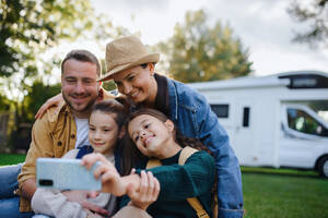 Eine fröhliche vierköpfige Familie hält einen Moment mit ihrem Wohnwagen im Hintergrund fest und lächelt für ein Selfie im Freien - HPIF07668