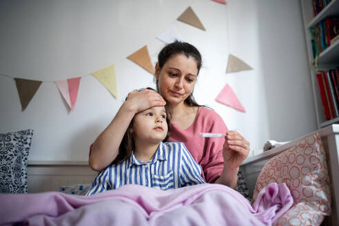 Portrait of mother looking after sick small daughter in bed at home, measuring temperature. - HPIF07687