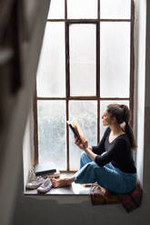 Portrait of happy woman sitting on old and dirty window sill, reading a book. - HPIF07721