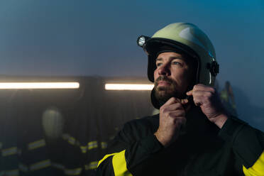 A mature firefighter preparing for action in fire station at night, looking at camera. - HPIF07734