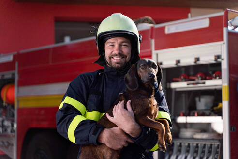 Ein glücklicher reifer Feuerwehrmann, der einen Hund hält und in die Kamera schaut, mit einem Feuerwehrauto im Hintergrund - HPIF07739