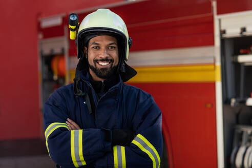 Ein glücklicher junger afroamerikanischer Feuerwehrmann mit Feuerwehrauto im Hintergrund. - HPIF07742