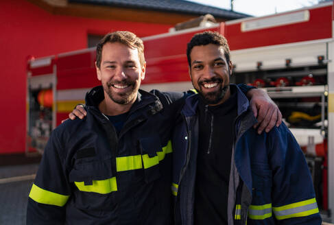 Glückliche Feuerwehrleute mit einer Feuerwache und einem Löschfahrzeug im Hintergrund, die in eine Kamera schauen. - HPIF07744