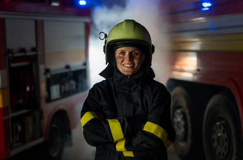 Ein mittlerer Erwachsener schmutzig weiblichen Feuerwehrmann Blick auf die Kamera mit Feuer LKW im Hintergrund in der Nacht. - HPIF07759