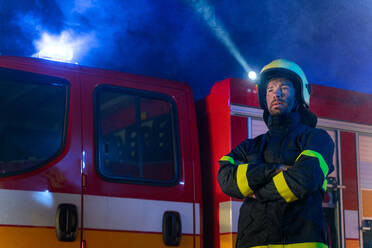 A low angle view of m firefighter talking to walkie talkie with fire truck in background at night. - HPIF07770
