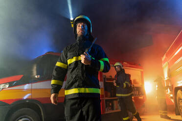 A low angle view of m firefighter talking to walkie talkie with fire truck in background at night. - HPIF07775