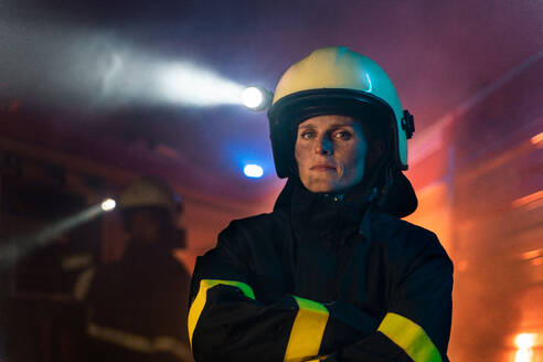 Ein schmutziger weiblicher Feuerwehrmann schaut in die Kamera mit einem Feuerwehrauto im Hintergrund bei Nacht. - HPIF07788