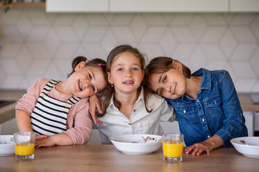 A portrait of three girls sisters indoors at home, looking at camera when eating breakfast. - HPIF07841