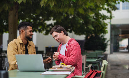 Ein junger Mann mit Down-Syndrom und sein betreuender Freund sitzen im Freien in einem Café und benutzen einen Laptop. - HPIF07854