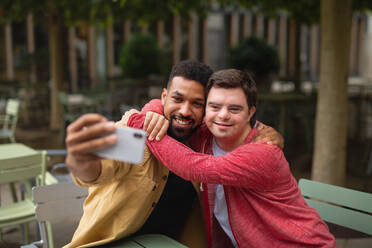 A young man with Down syndrome and his mentor enjoy a selfie moment while sitting outdoors at a cafe - HPIF07862