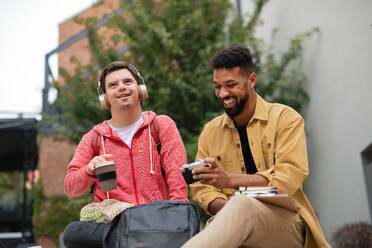 Ein junger Student mit Down-Syndrom und sein betreuender Freund sitzen im Freien auf dem Campusgelände - HPIF07873