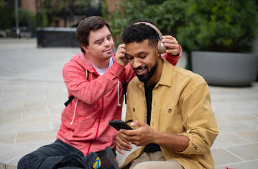 Ein junger Mann mit Down-Syndrom und sein betreuender Freund sitzen im Freien und hören Musik. - HPIF07874