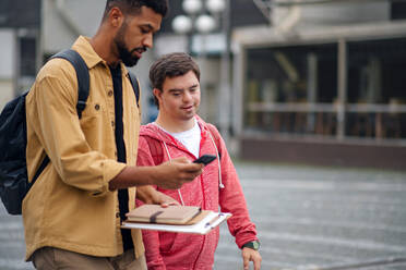 Ein junger Mann mit Down-Syndrom und sein betreuender Freund mit Smartphone gehen und sprechen im Freien - HPIF07878