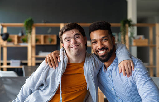 Happy young man with Down syndrome and his tutor with arms around looking at camera indoors at school - HPIF07895