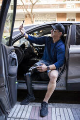 Smiling man with prosthetic leg taking selfie through smart phone sitting in car - JJF00336