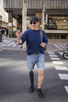 Lächelnder Mann mit Beinprothese macht Selfie und überquert Straße - JJF00339