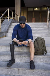 Älterer Mann mit Behinderung, der auf einer Treppe sitzend ein Smartphone benutzt - JJF00346