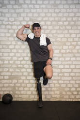 Glücklicher Mann mit Beinprothese, der seine Muskeln in der Nähe der Wand im Fitnessstudio anspannt - JJF00362