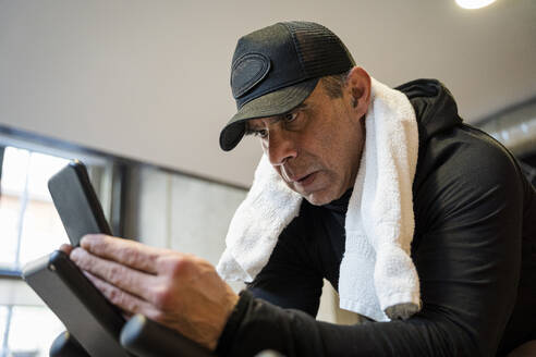 Konzentrierter Mann mit Mütze und Smartphone im Fitnessstudio - JJF00372