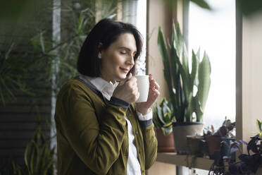 Woman smelling coffee standing by plants - NJAF00263