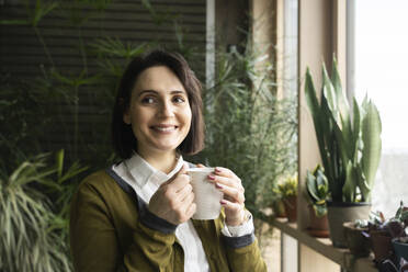 Smiling woman holding coffee cup in front of plants - NJAF00264