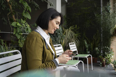 Smiling woman using smart phone sitting on chair by plants - NJAF00266