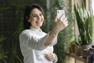 Glückliche Frau nimmt Selfie durch Smartphone vor Pflanzen - NJAF00267