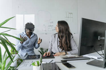 Colleague wearing virtual reality headset discussing with architect sitting at desk in office - OSF01404