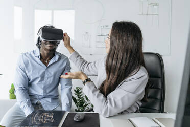 Architect adjusting virtual reality headset for colleague sitting at desk in office - OSF01405