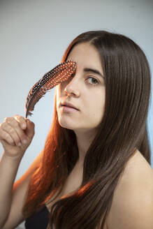 Young woman touching face with feather in front of wall - AXHF00342