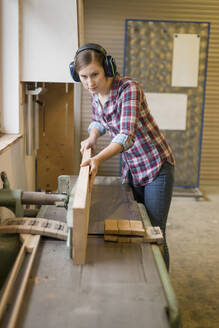 Craftswoman wearing ear protectors cutting wood in workshop - PAF01978