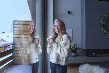 Glückliche Frau mit Kaffeetasse am Fenster stehend - JAHF00180