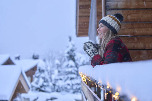 Happy woman with coffee cup enjoying snow in balcony - JAHF00192