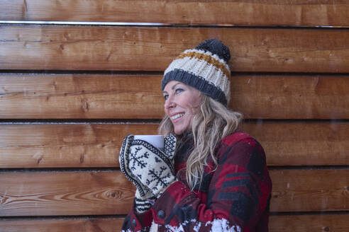 Happy mature woman with coffee cup leaning on wooden wall - JAHF00195