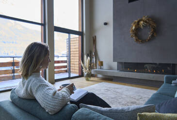 Woman sitting with book and coffee cup sitting on chair at home - JAHF00293